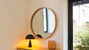 Una habitación con un armario de madera, un espejo redondo, una lámpara y vistas a un jardín verde a través de una puerta de vidrio.