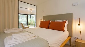 Een net opgemaakt bed met wit en oranje beddengoed, een houten nachtkastje en een raam met uitzicht op groen.
