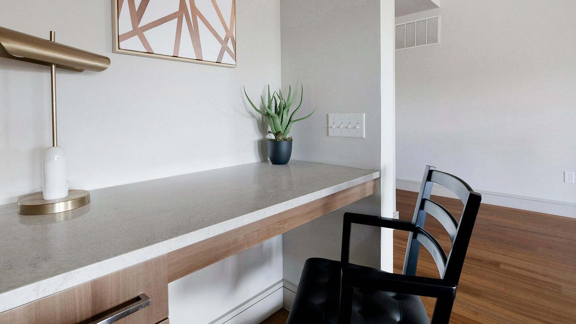 A modern kitchen with a wooden countertop, a black chair, a potted plant, and a white wall with a geometric artwork.
