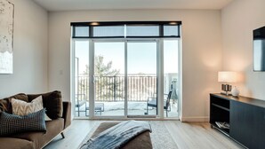 A modern living room with a sofa, a bench, a TV, and a balcony with outdoor furniture. - Belmont Properties Short-Term Rentals (Gatineau)