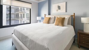 Een slaapkamer met een groot bed, twee kussens, een nachtkastje met een lamp, een raam met uitzicht op gebouwen en een aan de muur hangend schilderij.