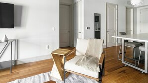 Een moderne woonkamer met een houten salontafel, een witte fauteuil, een eettafel met barkrukken, en een flatscreen televisie die aan de muur is bevestigd.