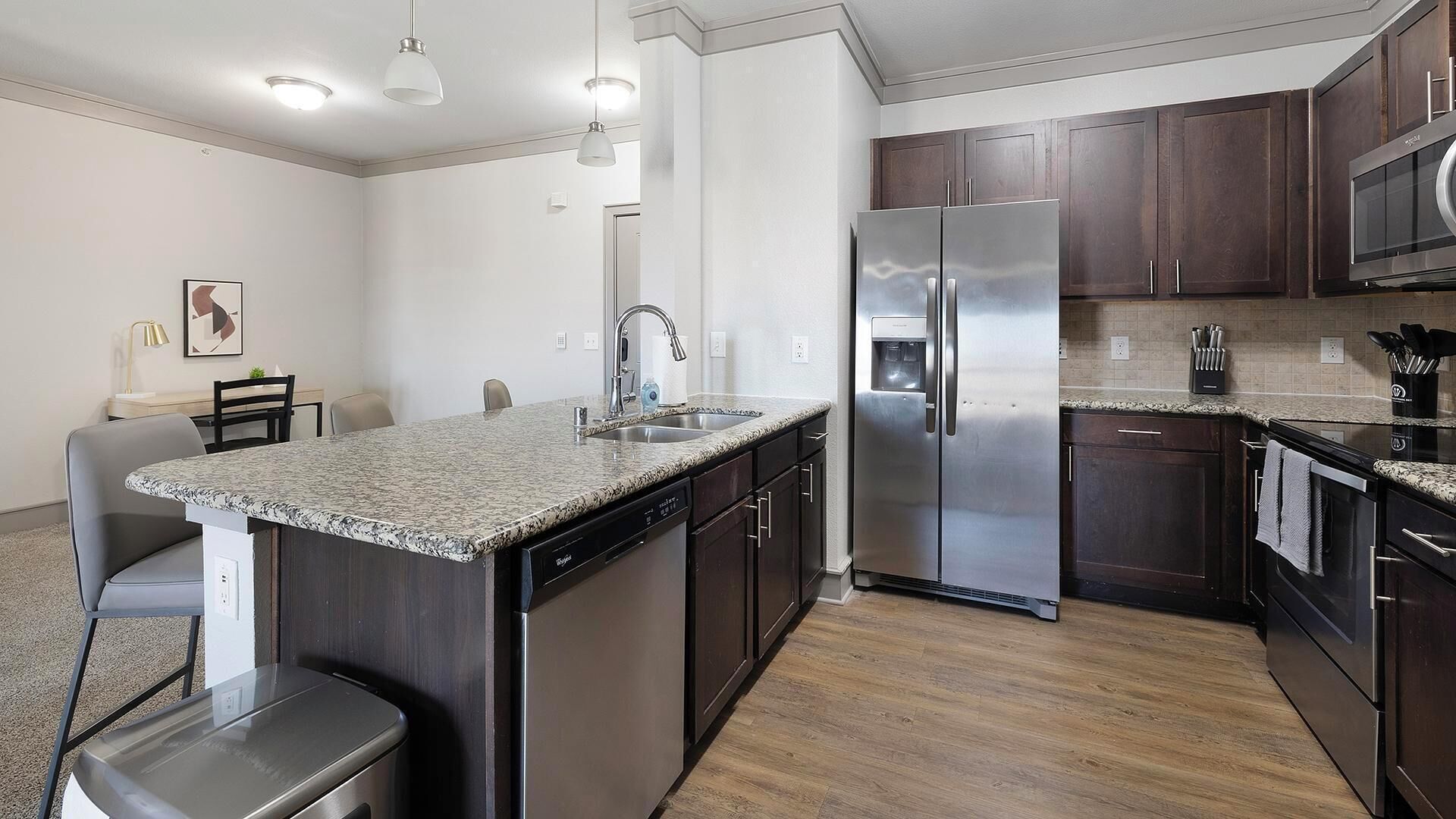 A modern kitchen with a granite countertop, stainless steel appliances, dark wood cabinets, and a dining area with a chair and table.