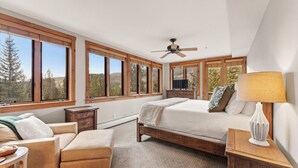 Un dormitorio con cama, una silla, una mesita de noche, una lámpara y ventanas amplias con vistas a árboles y colinas.