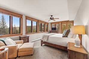 A bedroom with a bed, a chair, a nightstand, a lamp, and large windows with a view of trees.