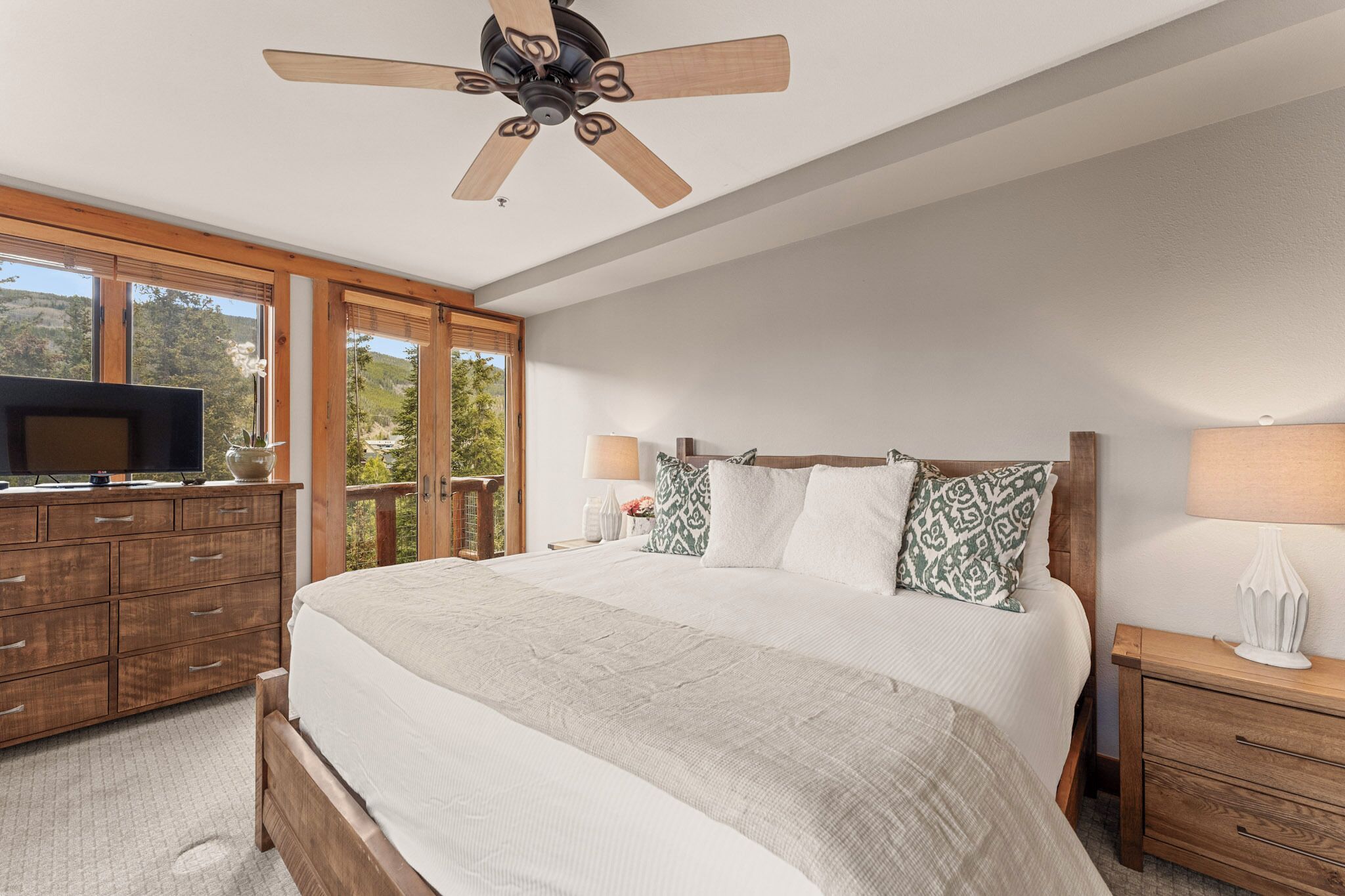 Un dormitorio con cama, cómoda de madera, televisor, ventilador de techo y ventanas grandes.