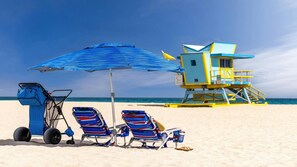 A beach scene with a lifeguard tower, two beach chairs, a blue umbrella, and a beach cart. - Miami Beach King Studio – Steps to Sand & Nightlife (Miami Beach)