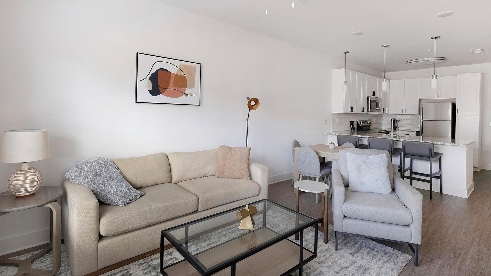 A modern living room with a sofa, armchair, coffee table, and a dining area with a table and chairs. A kitchen with white cabinets and appliances is visible in the background.