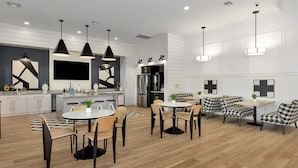 Une cuisine moderne avec des meubles blancs, un îlot central et des luminaires suspendus. On y trouve un coin repas avec une table ronde et des chaises, ainsi qu’un coin salon avec un canapé et des fauteuils à motifs à carreaux.
