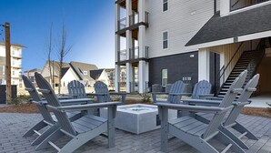Un espace extérieur aménagé en terrasse, avec des fauteuils Adirondack et un foyer central, le tout entouré de bâtiments modernes.