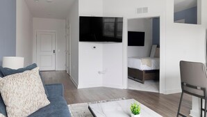 Una sala moderna con un sofá, una mesita de centro con una planta, un televisor montado en la pared y una cama en la habitación contigua.