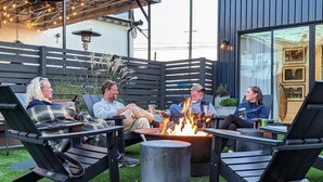 Eine Gruppe von Menschen sitzt um eine Feuerstelle auf einer Terrassenanlage, im Hintergrund ein modernes Gebäude.