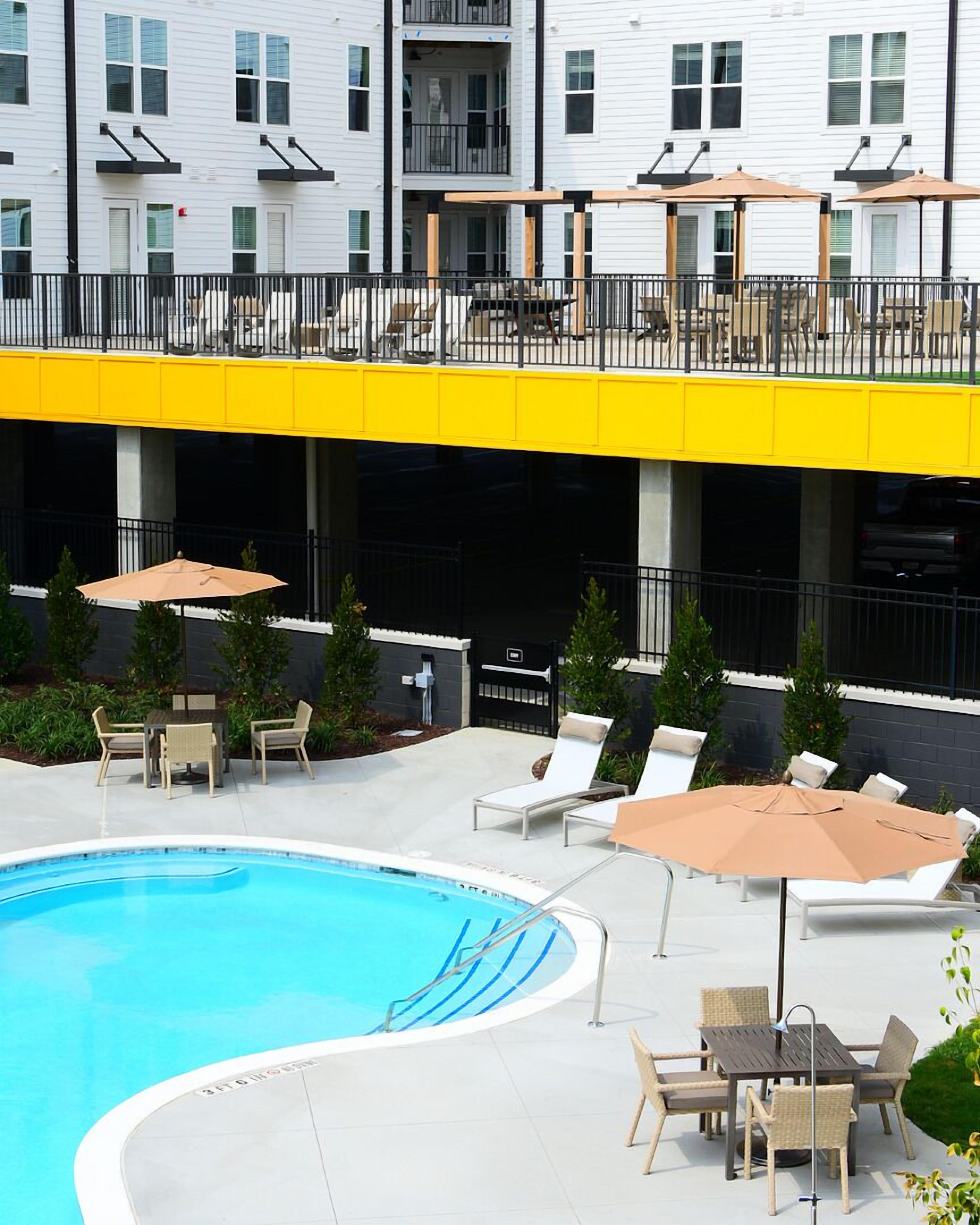 Un área de piscina con sillones de descanso y sombrillas, una terraza techada y un edificio con múltiples ventanas y balcones.