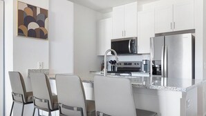 Una cucina moderna con mobili bianchi, un frigorifero in acciaio inossidabile, un forno a microonde e una macchina da caffè. Sono presenti tre sedie e un piano di lavoro in marmo.