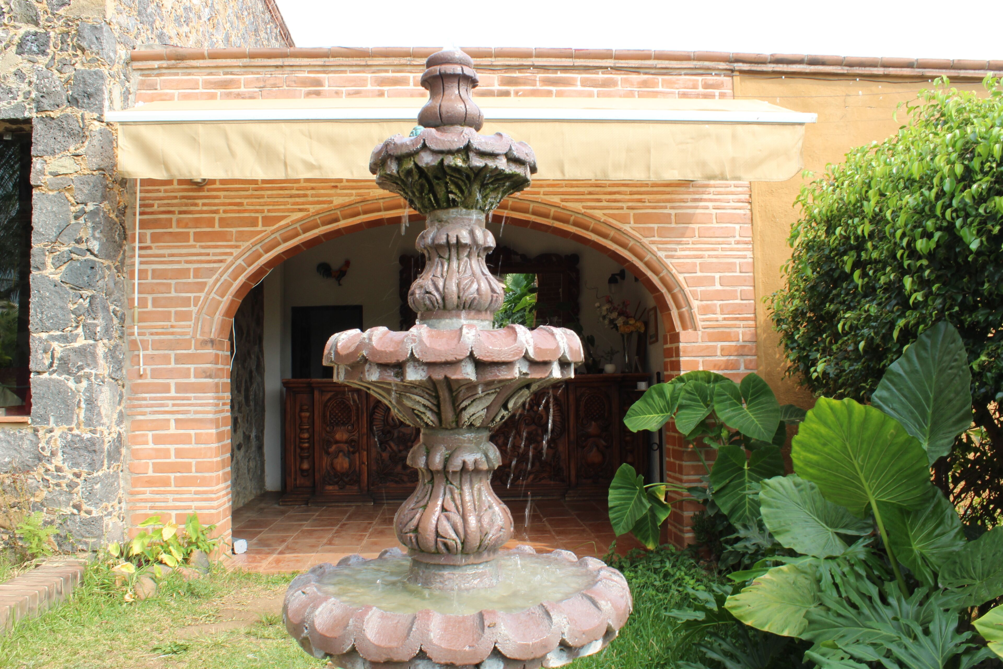 A stone fountain with multiple tiers, a brick building with an arched entrance, and large green foliage in the foreground.