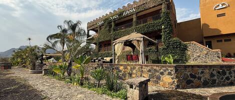 A building with a sign reading 'Casa Juana,' featuring a stone facade, a covered outdoor seating area, and surrounding greenery.