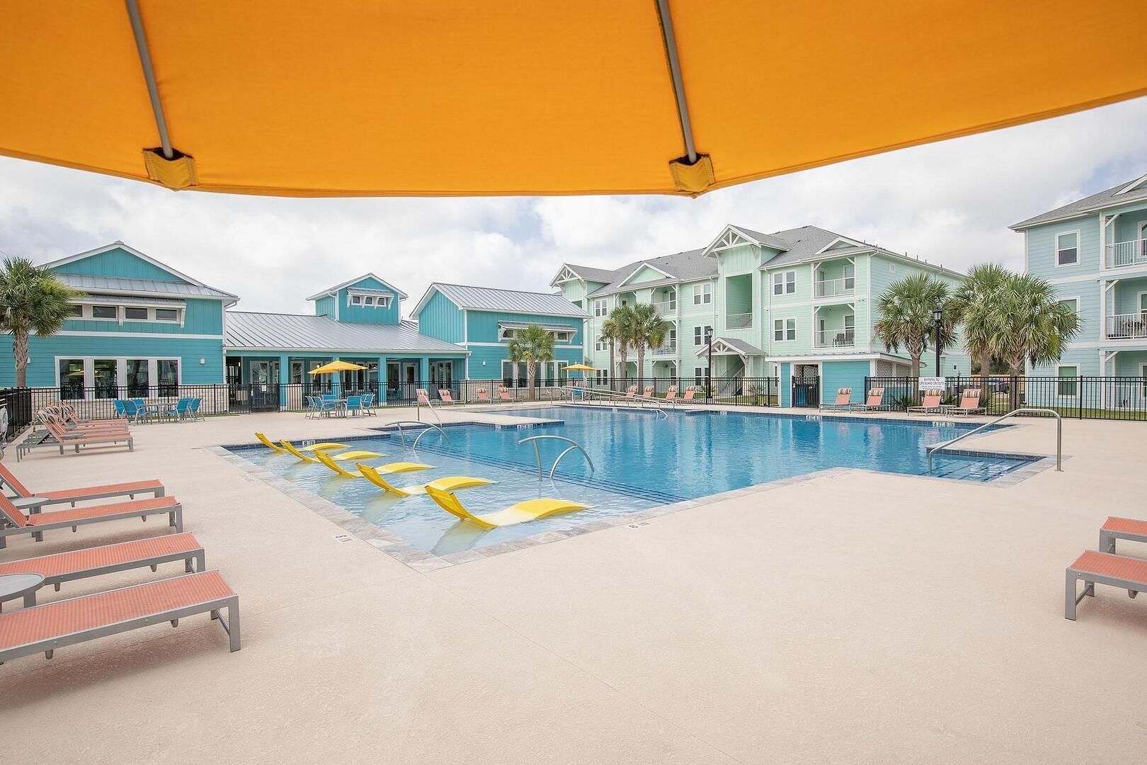 A swimming pool area with lounge chairs and umbrellas, surrounded by buildings with a light blue facade.
