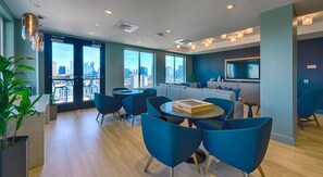 A modern hotel room with large windows, a city view, a dining area with blue chairs, and a flat-screen TV mounted on the wall. - Landing | Beautiful 1BD, Pool, Gym (Nashville)