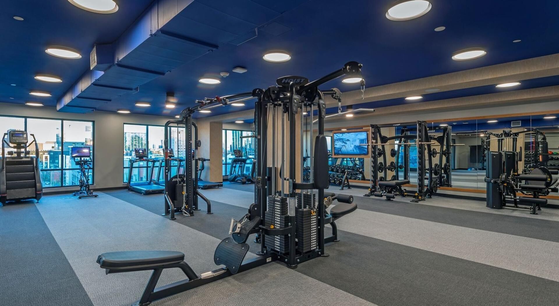 Une salle de sport moderne, dotée de divers appareils d’exercice, de grandes fenêtres et offrant une vue sur le paysage urbain.
