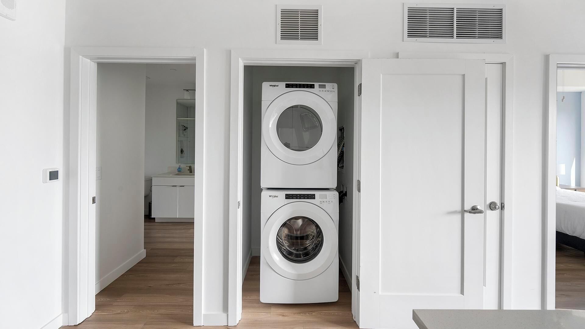 A laundry room with two stacked washers and dryers, a door to a bathroom, and a hallway leading to a bedroom.