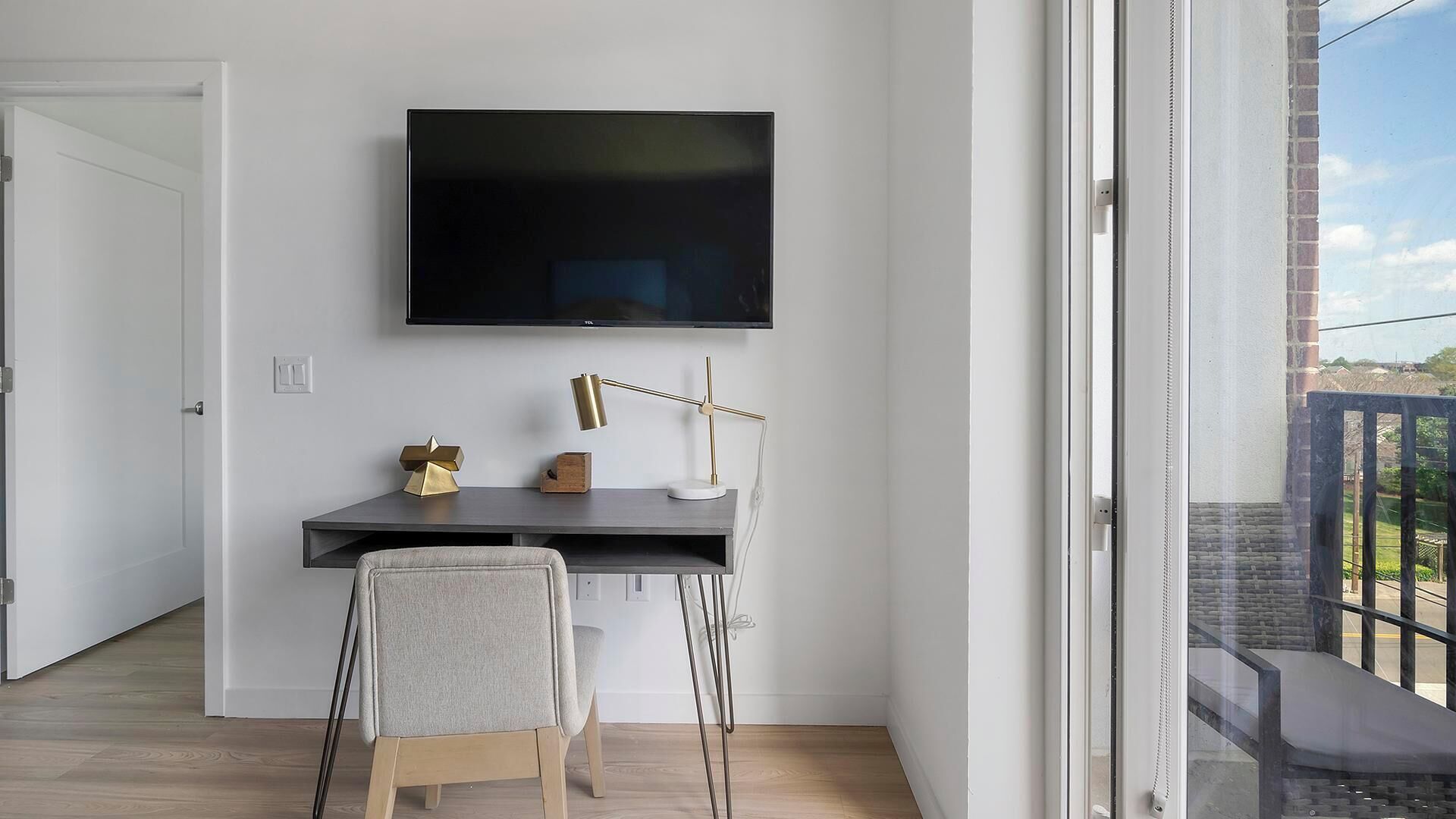 Una camera moderna con una TV a schermo piatto montata a parete, una scrivania con una lampada, una sedia e vista sull'esterno da una grande finestra.