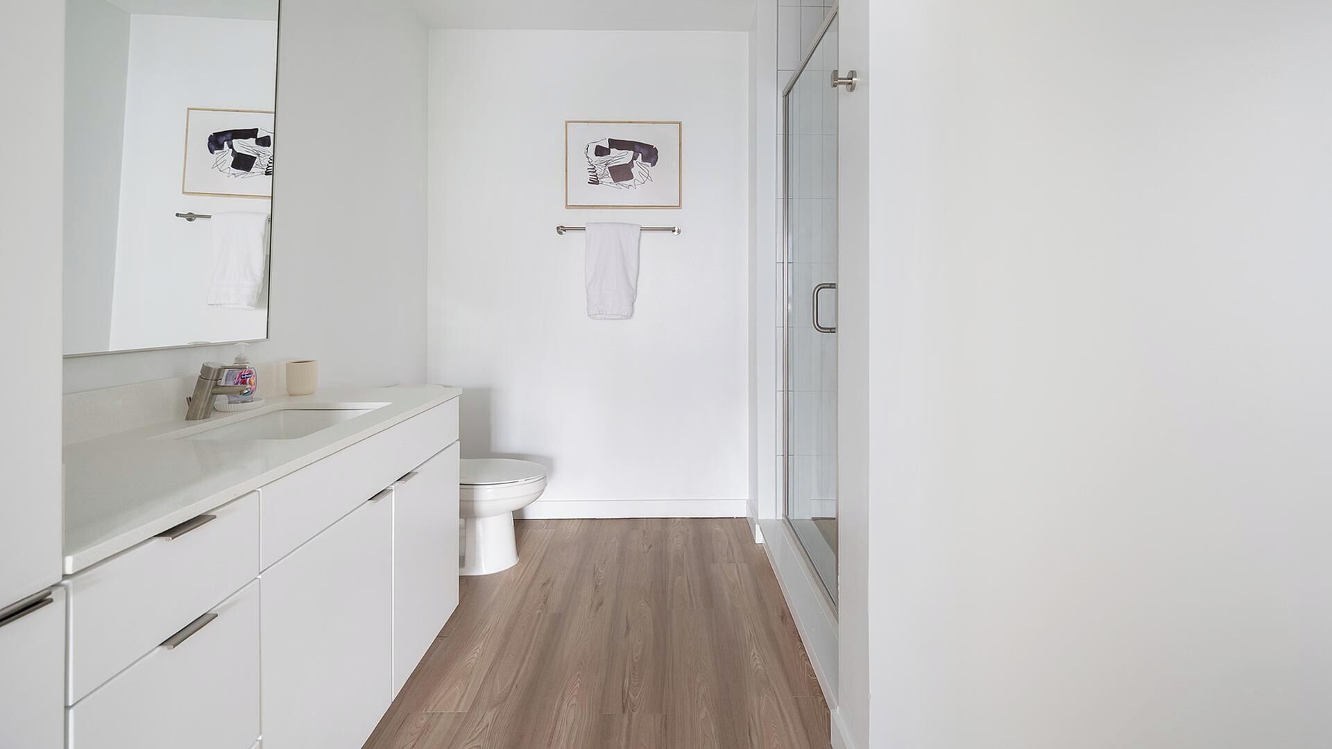 A modern bathroom with a white sink, mirror, and toilet, wooden flooring, and two framed pictures on the wall.