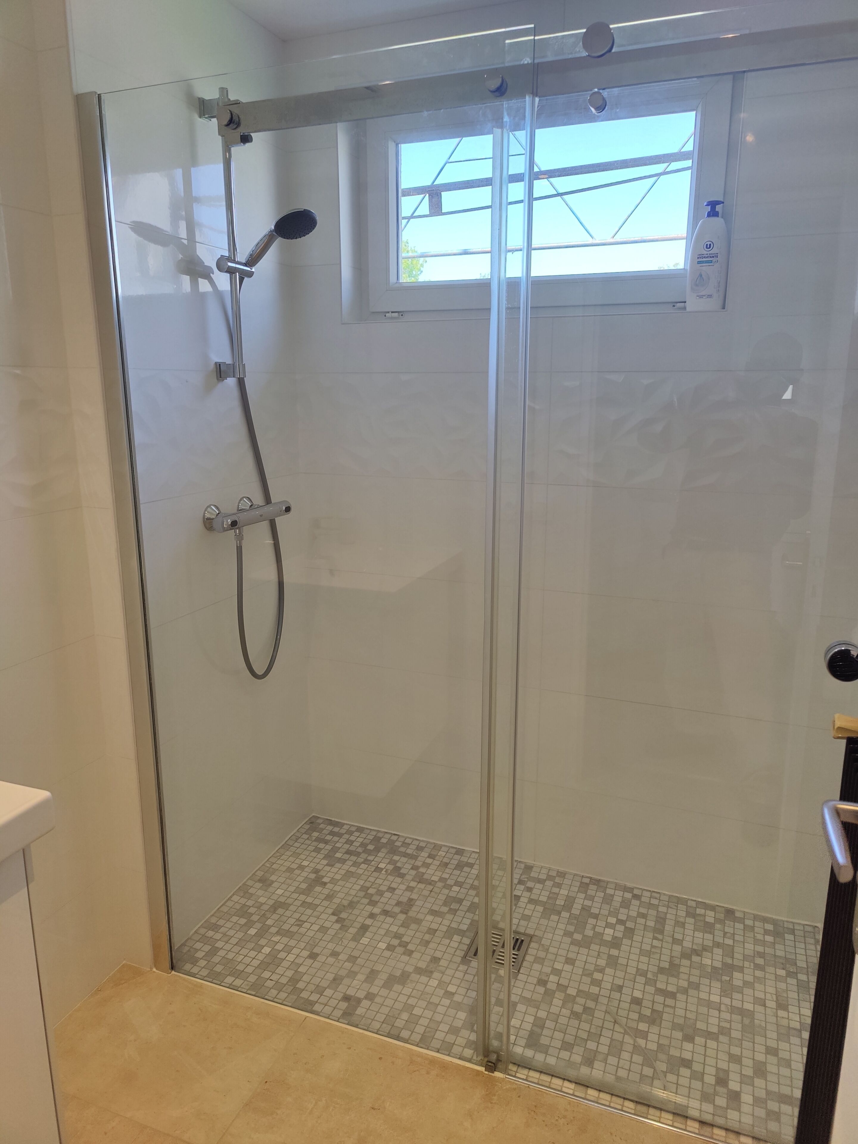 Ducha con cerramiento de vidrio, ventana, suelo de baldosas y dispensador de jabón a la vista.