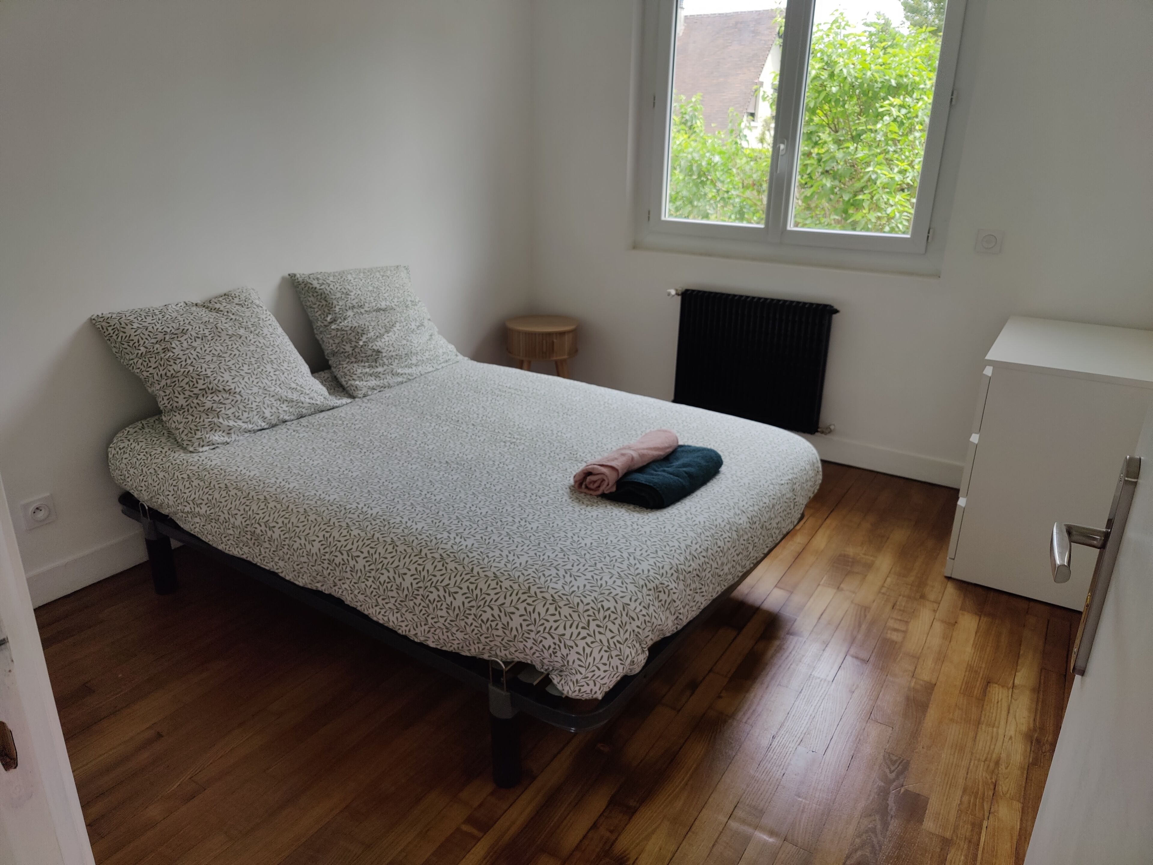 A bedroom with a bed, a radiator, a small wooden table, and a white cabinet.