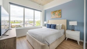 Un dormitorio con una ventana grande, una cama, una mesita de noche y un cuadro en la pared.