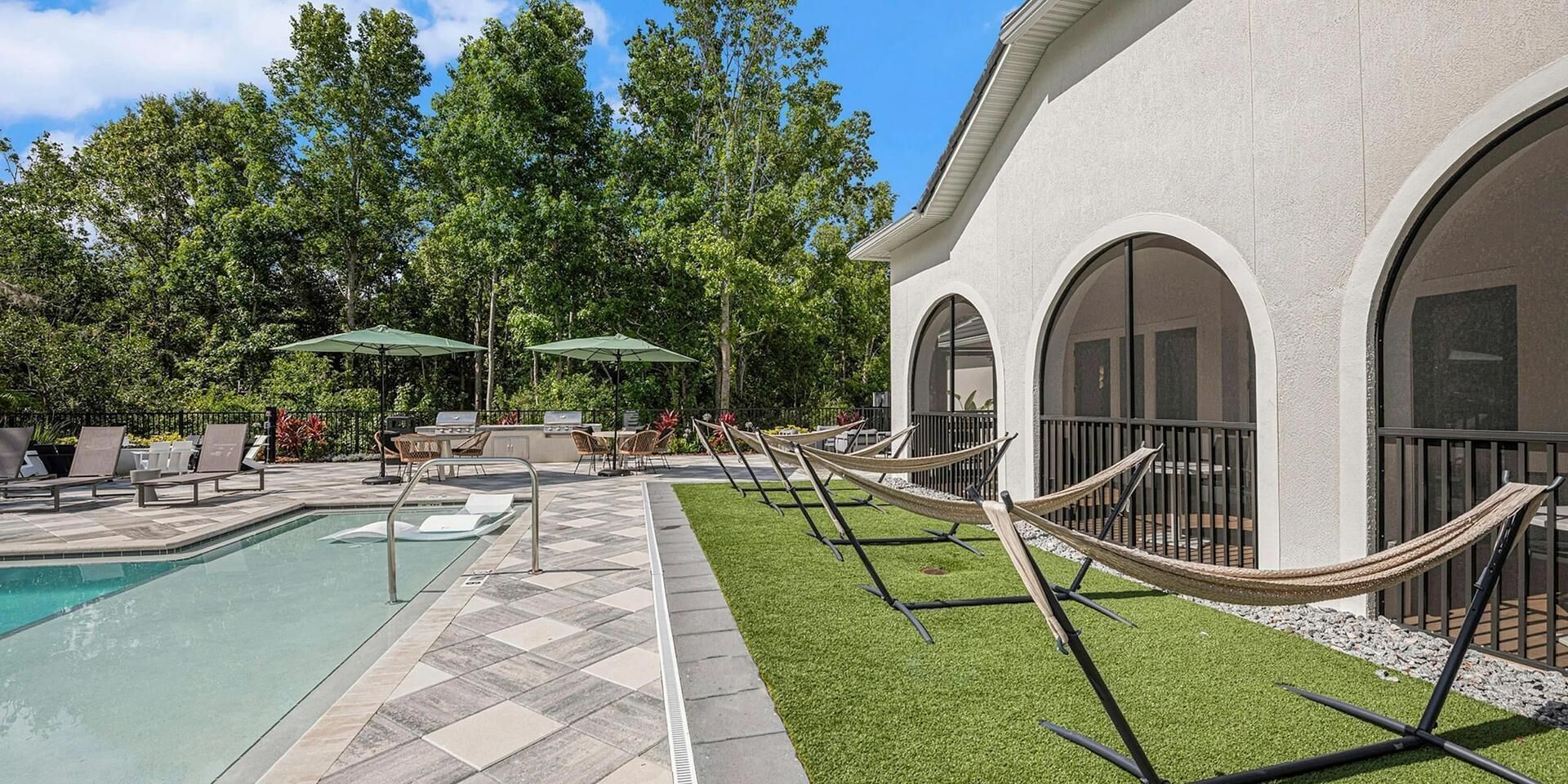 A pool area with a tiled deck, lounge chairs, a hammock, and a building with arched windows.