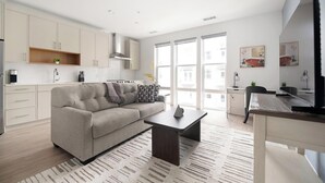 A modern living room with a sofa, coffee table, and a dining area with a chair and table. - Landing | Amazing 1BD, Pool, Gym (Hyattsville)