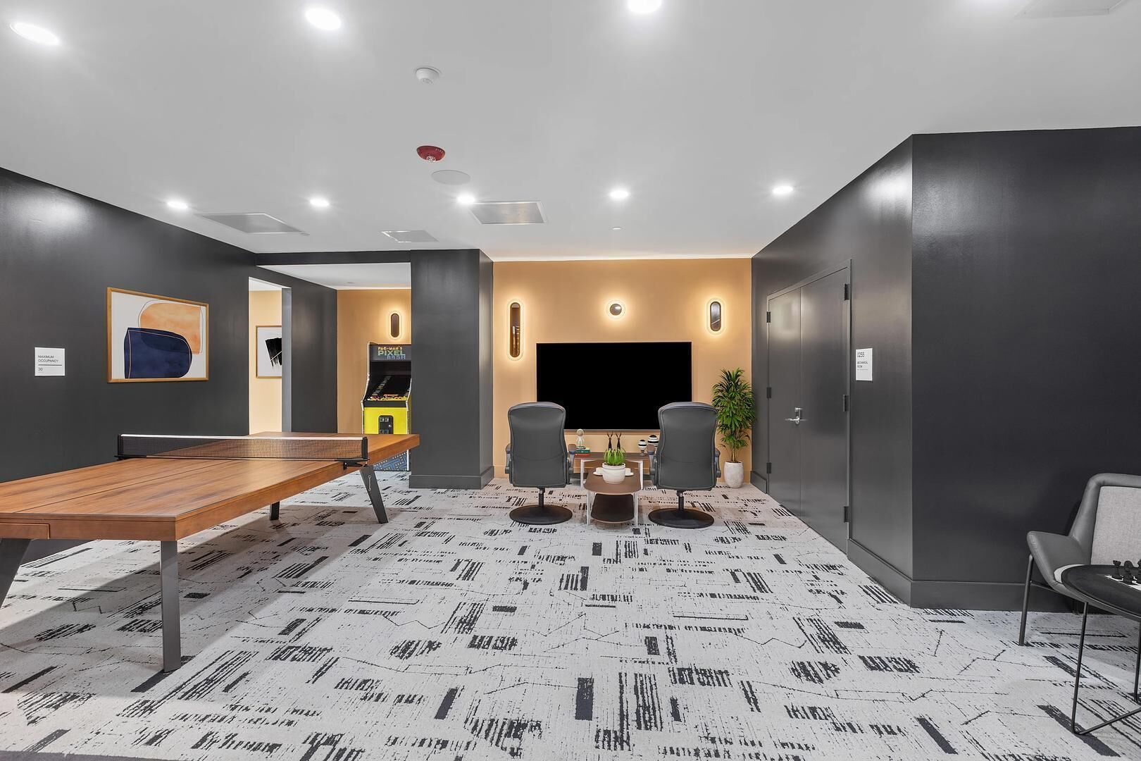 A modern conference room with a long table, two chairs, a TV, and a gaming machine.