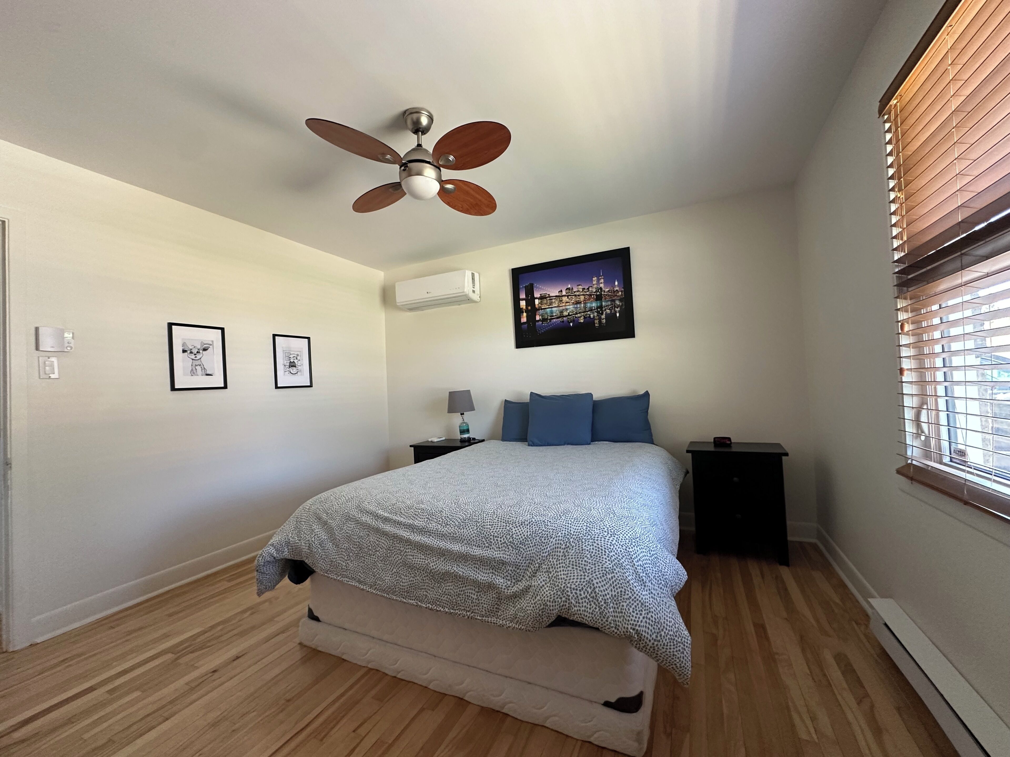 Ein Schlafzimmer mit einem Bett, einem Deckenventilator, einem Nachttisch, einer Lampe, einem Bilderrahmen und einem Fenster mit Jalousien.