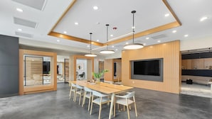 A modern conference room with a long table, white chairs, pendant lighting, a large flat-screen TV, and wooden paneling on the walls. - Landing | Chic 1BD, Gym, Game Room (Hyattsville)