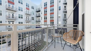 A balcony with a view of a multi-story building, a small table, and two wicker chairs. - Landing | Chic 1BD, Gym, Game Room (Hyattsville)