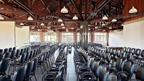 Uma sala de conferências com fileiras de cadeiras pretas, vigas de madeira no teto e janelas amplas.
