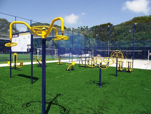 A playground with yellow and blue exercise equipment, a fenced area, and a grassy field. - POUSADA FECEP (Guaratuba)