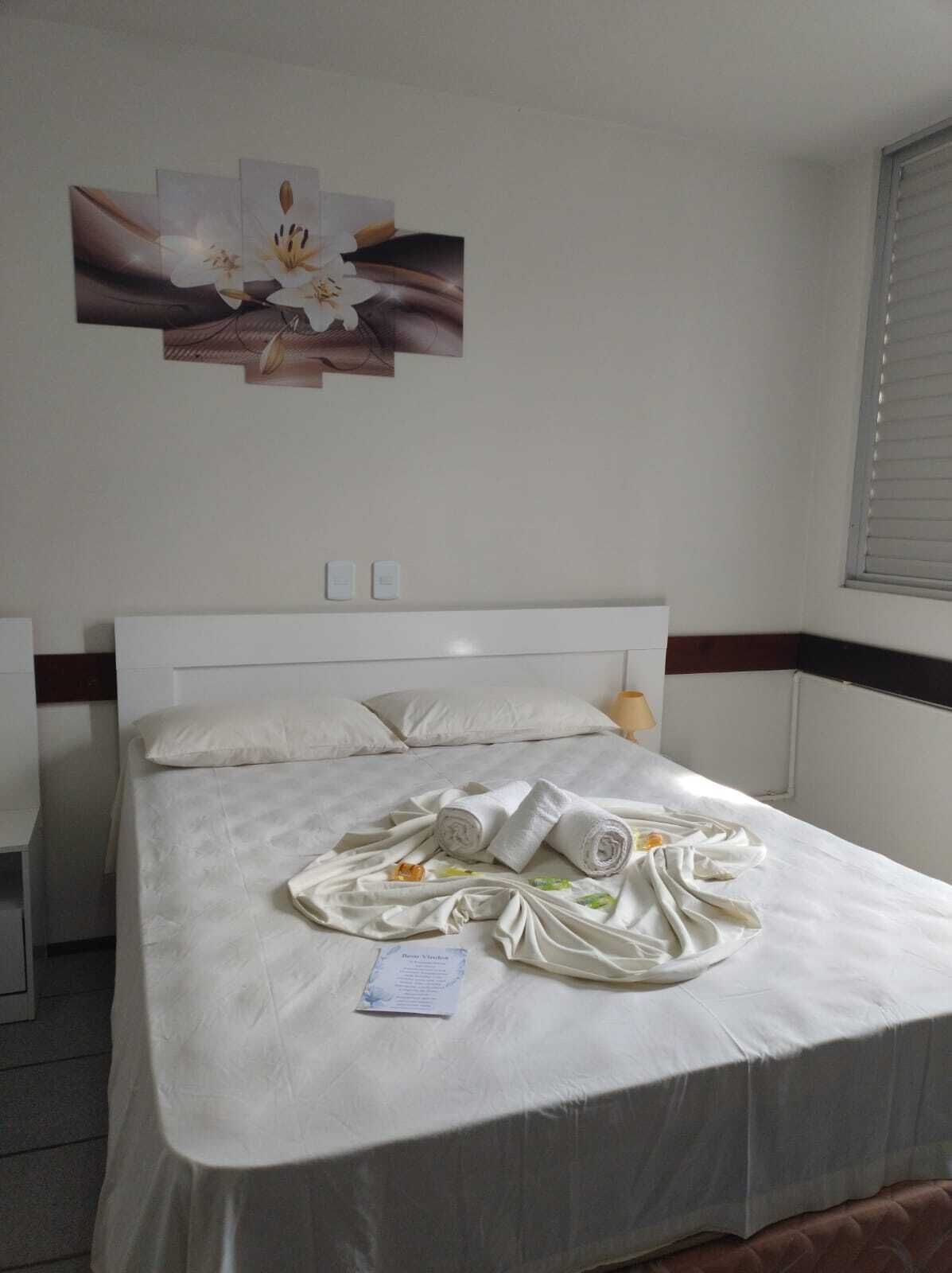 A hotel room with a neatly made bed, a painting on the wall, and a window with blinds.