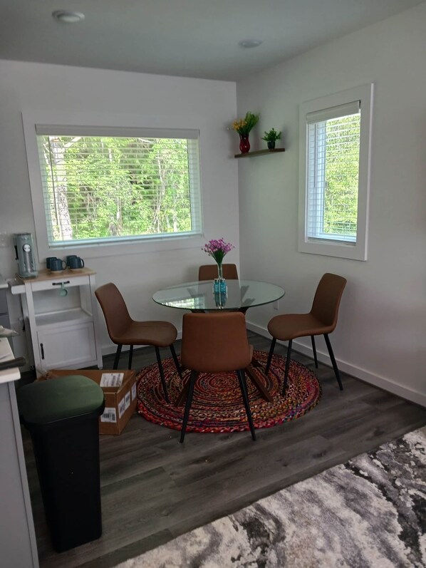 A dining area with a glass table, four chairs, a vase with flowers, and two windows with blinds.
