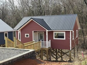 A red cabin with a wooden deck and a metal roof, elevated on stilts, surrounded by trees.