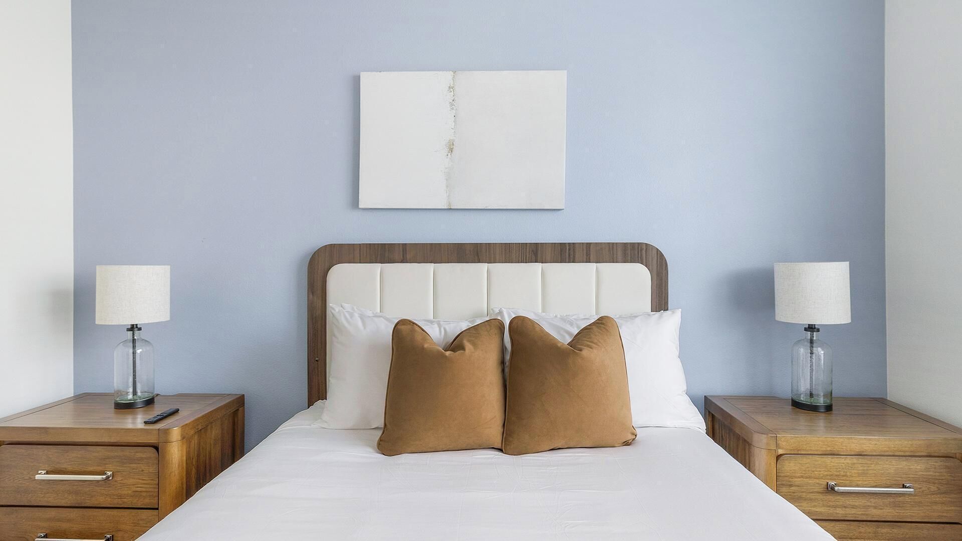 Ein ordentlich bezogenes Bett mit zwei Nachttischen, jeweils mit einer Lampe, in einem Zimmer mit hellblauer Wand und einem abstrakten Kunstwerk darüber.