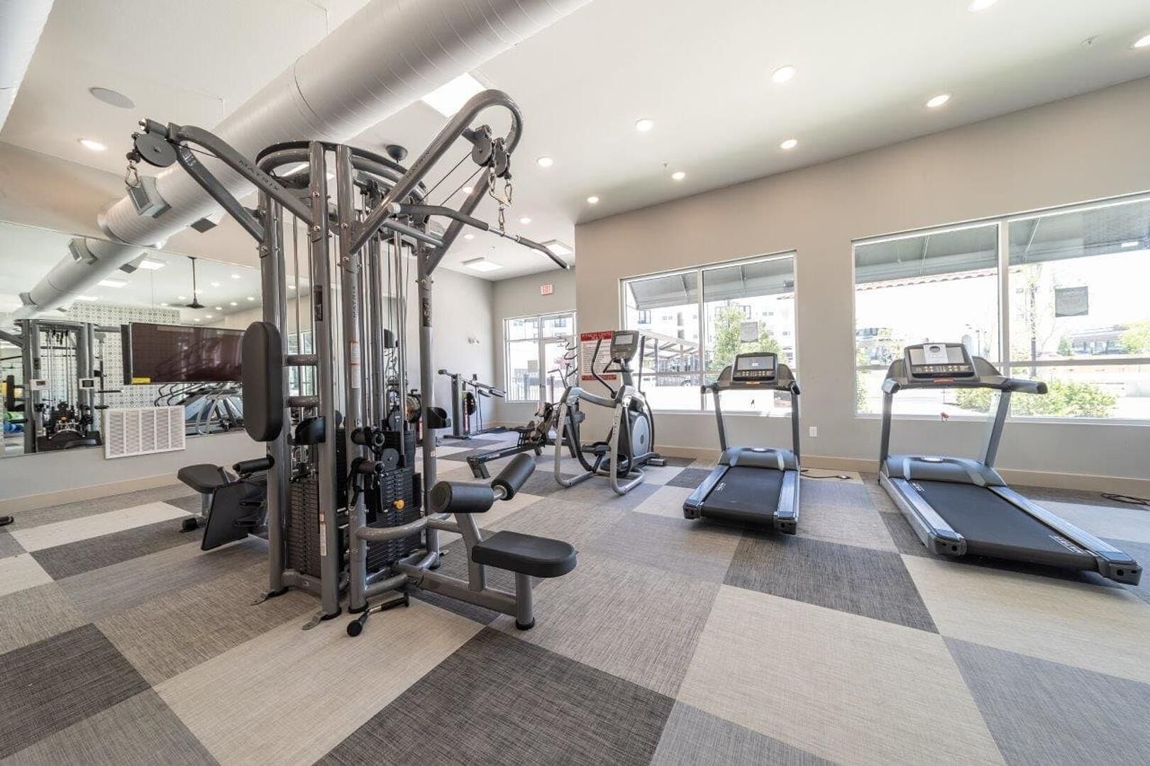 A gym with cardio machines, weight training equipment, and a large window providing natural light.
