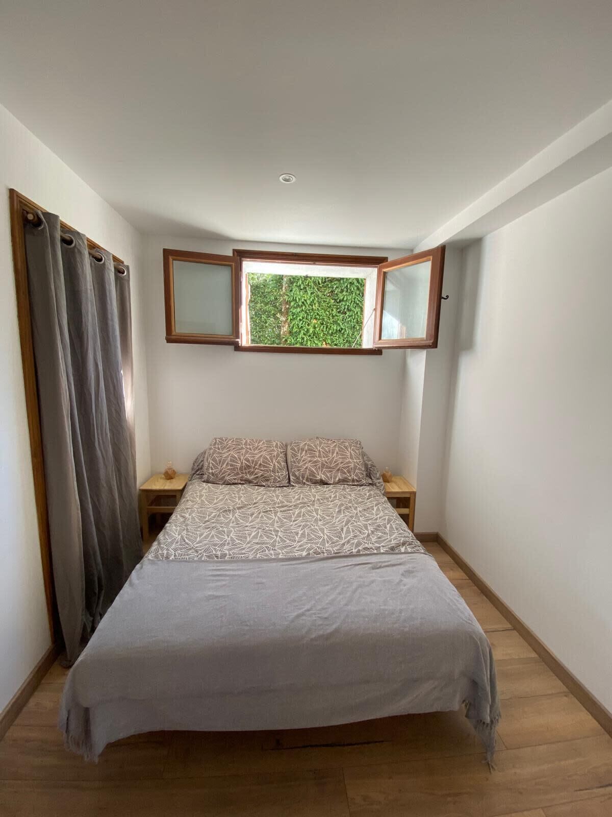A bedroom with a bed, a window with a view of greenery, a nightstand, and a curtain.