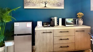 A kitchenette with a refrigerator, microwave, and coffee maker on a wooden cabinet, a painting of a tree on the wall, and a potted plant.