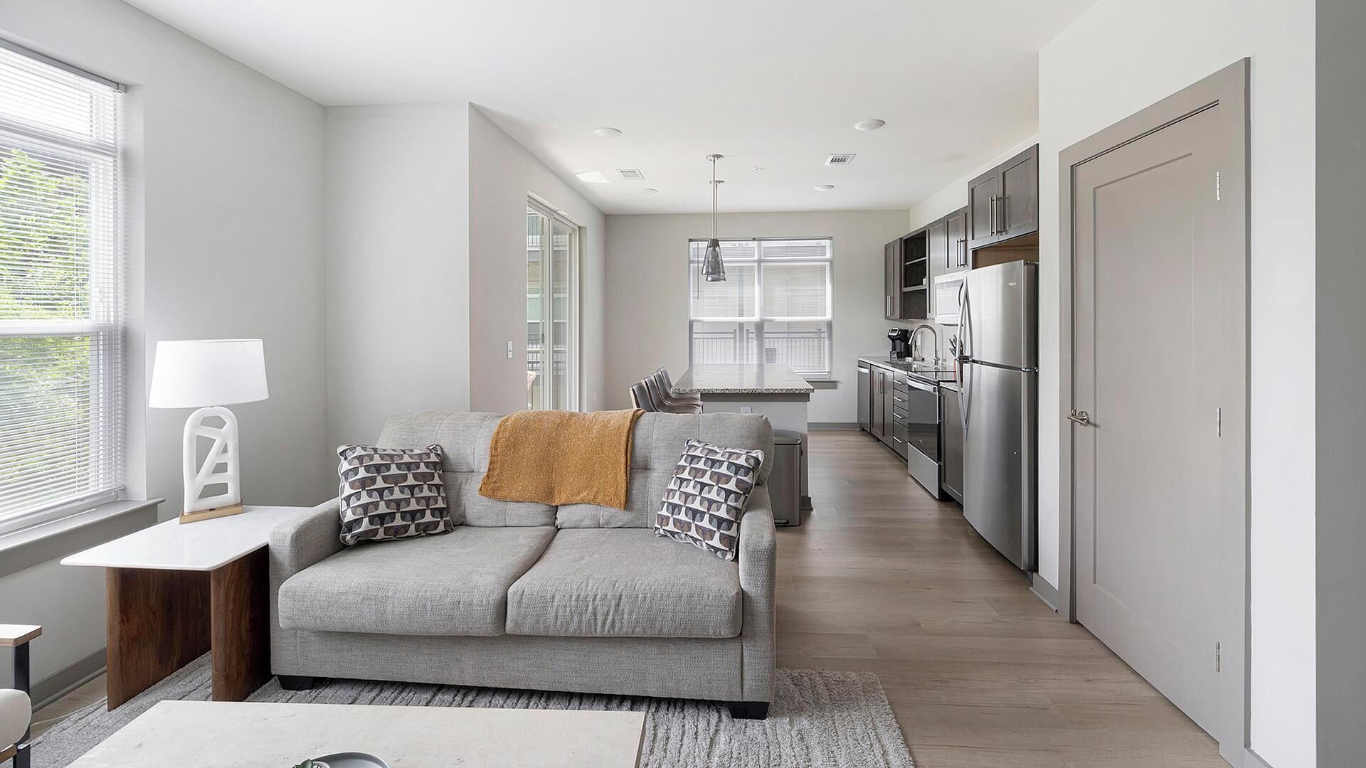 A modern kitchen with stainless steel appliances, a grey sofa with patterned cushions, and a window with blinds.