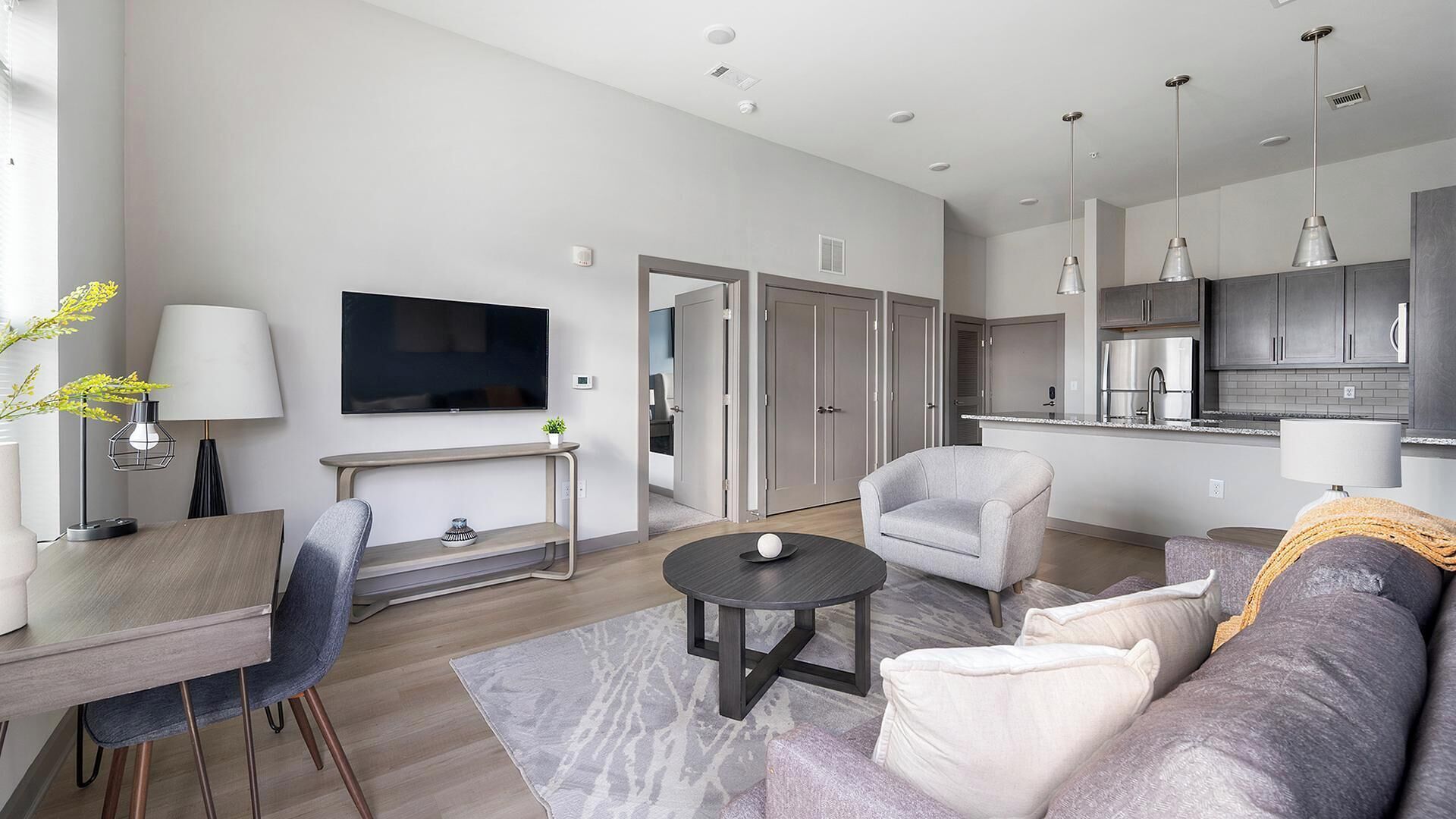 A modern living room with a flat-screen TV, a dining table, a sofa, and a kitchen area with cabinets and a refrigerator.
