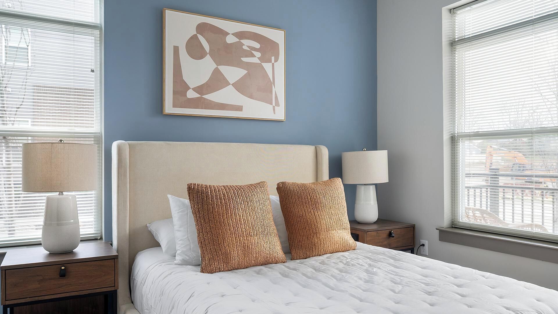A bedroom with a bed, two pillows, a nightstand, a lamp, a wall art piece, and a window with blinds.