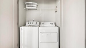 Une buanderie équipée de deux machines à laver et de deux sèche-linge Whirlpool blancs, d’un panier à linge blanc posé sur une étagère et d’un mur blanc.