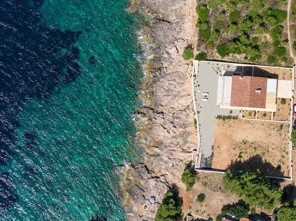 An aerial view of a coastal property with a building adjacent to the shoreline, a paved area, and sparse vegetation. - Villa Lyra by Villas Hvar (Ivan Dolac)