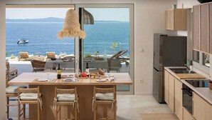 Une cuisine avec vue sur la mer, un coin repas avec des chaises et un grand îlot central surmonté d’un luminaire suspendu.